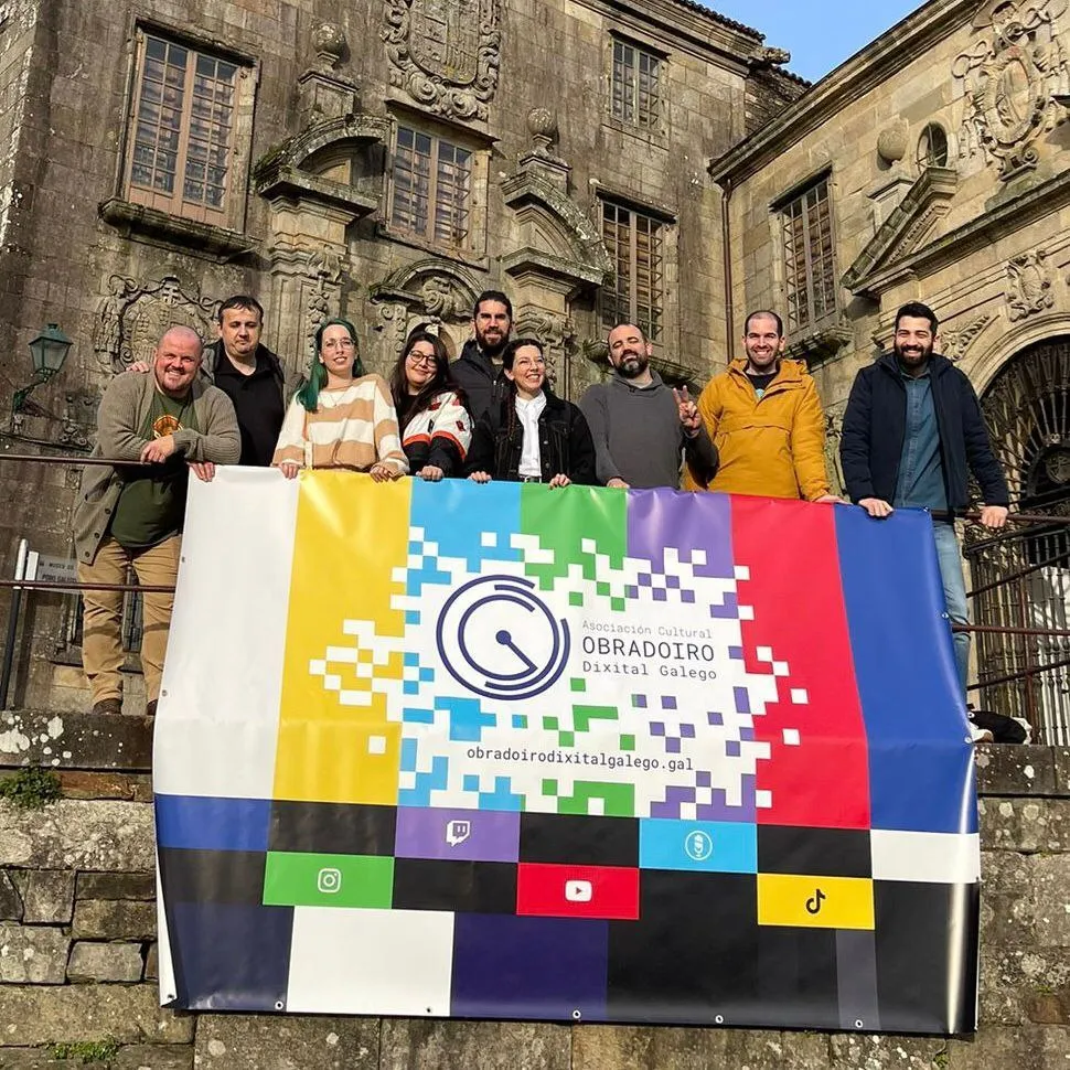 Fotografía no exterior do Museo do Pobo Galego coa faixa co cartón de axuste de Obradoiro Dixital Galego. Detrás dela e de esquerda a dereita están Fran da Lobeira, David Paz (Madialevo), Laura Veiga (Pingando), Sofía Naseiro (Kinocube Cinema),  Sergio e Nuria (Toxío), Miguel González (Alkaine), Pablo Villaverde (Fedello do Clan Kirfed) e Carlos Vieito (Exército Mekemeke)